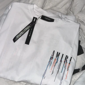 Vit AMIRI t-shirt med färgstänk - Säljer en vit t-shirt från AMIRI med coolt färgstänksmönster och logga på bröstet. T-shirten har rund hals och korta ärmar, perfekt för dig som gillar streetwear med unik design. Materialet är mjuk bomull och passformen är normal.