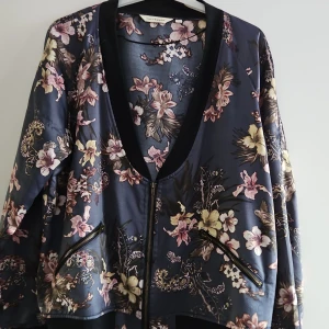 Blommig bomberjacka  - Snygg bomberjacka i mörkblått med blommigt mönster i rosa, beige och gult. Jackan har dragkedja framtill, svarta detaljer vid halsringningen och två snedställda fickor med dragkedja. Perfekt för dig som inte använder kavajer 