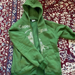 Grön hoodie med tryck Capo Regim - Säljer en grön hoodie från Capo Regim med dragkedja och huva. Framsidan har ett stort tryck med text och motiv av en figur med lie. Hoodien har kängurufickor och ribbade muddar. Perfekt för dig som gillar streetwear vibes.