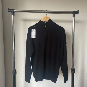 Svart half zip stickad tröja Stone Island - Svart stickad tröja från Stone Island med half zip och hög krage. Klassisk patch med logga på vänster ärm. Långärmad och ribbad vid muddar och nederkant. Perfekt för dig som gillar stilrena och snygga plagg med street-vibe.