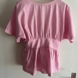 Rosa och vit randig blus med knyt - Säljer en söt blus med rosa och vita ränder, korta vida ärmar och knyt i midjan. Toppen har en avslappnad passform och markerad midja med volang nedtill. Ä