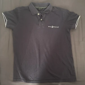 Mörkblå Sail Racing pikétröja i storlek S - Mörkblå pikétröja från Sail Racing i storlek S. Klassisk krage med två knappar och vit logga broderad på bröstet. Ärmarna har vit kantdetalj och materialet är mjuk bomull. Perfekt för dig som gillar sportig och stilren look.