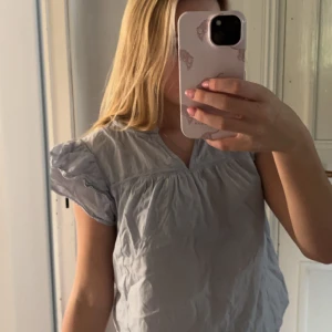 Ljusblå blus med puffärm - Säljer en ljusblå blus med korta puffärmar och v-ringning. Blusen har en lös passform och är tillverkad i ett tunt, lätt material som ger en luftig känsla. Perfekt för dig som gillar en enkel men söt stil.