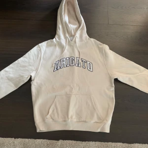 Beige Arigato hoodie med tryck - Snygg beige hoodie från Arigato med stor broderad logga på bröstet. Klassisk känguruficka, huva med snörning och mjukt bomullsmaterial. Perfekt för chill dagar eller när du vill ha en clean och stilren look.