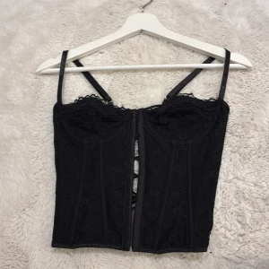 Svart spetskorsett från SHEIN - Svart korsett från SHEIN med smala axelband, spetsdetaljer och snörning i ryggen. Toppen har en figurnära passform och dekorativa sömmar framtill. Perfekt för dig som vill ha en edgy och trendig look.