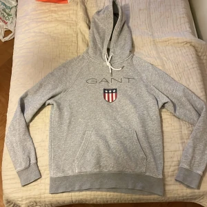 Grå hoodie från GANT  - Snygg grå hoodie från GANT med broderad logga och sköld på bröstet. Tröjan har huva med snörning, känguruficka och långa ärmar. Skulle säga storlek är mer S/M