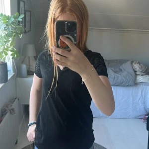 Svart topp med spetsdetaljer - Svart topp med v-ringning och korta ärmar i spets. Toppen har en enkel, rak passform och är tillverkad i mjukt material. Spetsdetaljer finns både vid halsringningen och på ärmarna vilket ger en snygg kontrast till resten av toppen.Den är har ändvänts max 4 gånger och är i fin kvalité.Den har inte hål eller fläckar 
