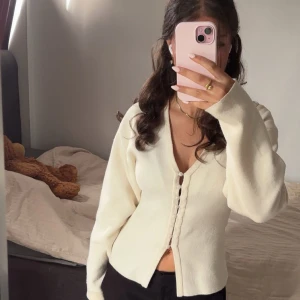 Krämvit kofta med v-ringning - Säljer en krämvit kofta med v-ringning och dekorativa knappar framtill. Koftan har långärmade ärmar och är tillverkad i ett mjukt, stickat material. Perfekt för lager-på-lager och enkel att styla till olika outfits.