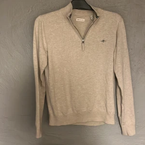 Beige half zip tröja från GANT - Snygg beige tröja från GANT med half zip och broderad logga på bröstet. Tröjan har lång ärm, ribbade muddar och en krage med svarta ränder på insidan. Ny köpt för 1000 SEK. Tillverkad i mjuk bomull, perfekt för lager-på-lager under höst och vår. 