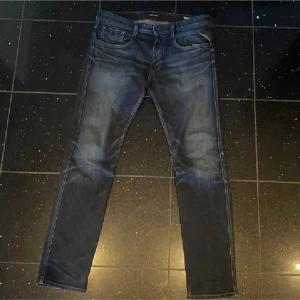 Replay Anbass jeans mörkblå, stl 32 - Säljer ett par Replay Anbass jeans i mörkblå tvätt med snygga slitningar och klassisk femficksdesign. Jeansen har raka ben och normal passform, med Replay-läderpatch bak i midjan. Perfekta för dig som gillar stilrena och bekväma jeans.