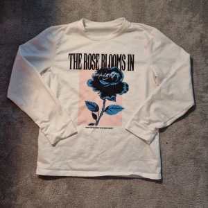 Vit långärmad tröja med blå ros från SHEIN - Vit långärmad tröja från SHEIN med tryck framtill som visar en blå ros och texten 'THE ROSE BLOOMS IN'. Tröjan har rund halsringning och är gjord i mjuk bomullsblandning. Perfekt för dig som gillar coola prints och vill sticka ut lite extra.