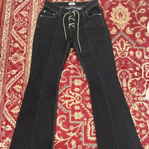 Svarta bootcut jeans med snörning - Snygga svarta jeans från Weekday med bootcut-modell och cool snörning framtill istället för vanlig dragkedja. Jeansen har klassiska fickor och markerade sömmar längs benen. Perfekta för dig som vill sticka ut lite extra.