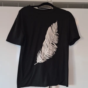 Svart t-shirt med vit fjädertryck - Cool svart t-shirt med ett stort vitt fjädertryck på framsidan. Klassisk rund hals och korta ärmar. Perfekt för dig som gillar stilrena och grafiska motiv. Materialet känns mjukt och bekvämt mot huden.