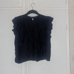 Svart blus med volang från Gina Tricot - Supersöt svart blus från Gina Tricot med volangdetaljer längs axlarna och spetskant vid halsen. Blusen är ärmlös och har en rak passform. Perfekt att styla med jeans eller kjol för en trendig look.