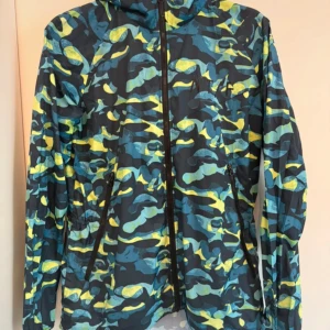 Nike vindjacka camo blå/gul L - Nike vindjacka i storlek L med blå, gul och svart camouflage-mönster. Jackan har huva, hel dragkedja framtill och två snedställda fickor med dragkedja. Tillverkad i lätt polyester, perfekt för blåsiga dagar. Snygg och sportig look.