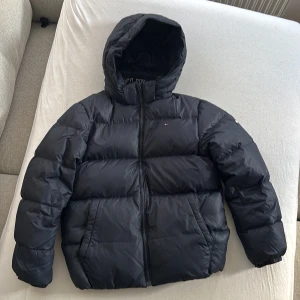 Mörkblå Pufferjacka Tommy Hilfiger - ❗️Kom gärna med prisförslag ❗️Mörkblå pufferjacka från Tommy Hilfiger med huva och dragkedja framtill. Jackan har broderad logga på bröstet och stora fickor på sidorna. Materialet är syntet och jackan är quiltad för extra värme. Perfekt för kalla dagar och har en clean, modern look.