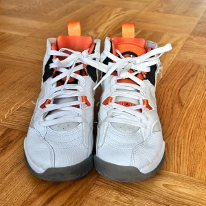Nike Air Jordan 6 sneakers vit/orange - Nike Air Jordan 6 sneakers i vitt skinn med orange och svarta detaljer. Höga med snörning, grå sula och tydlig Jumpman-logga på hälen. Orange dragflikar och meshpaneler på sidorna ger en sportig vibe. Perfekta för dig som vill sticka ut med klassisk Jordan-stil.