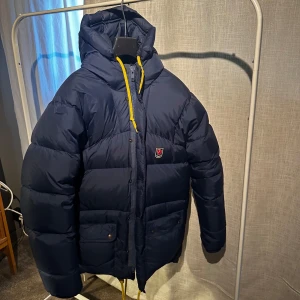 Mörkblå dunjacka från Fjällräven - Säljer en mörkblå dunjacka från Fjällräven med gul dragsko och huva. Jackan har långa ärmar och praktiska fickor med knappar. Sitter stort. 