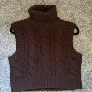 Brun stickad väst med polokrage - Mysig brun stickad väst med grovt kabelstickat mönster och hög polokrage. Västen är ärmlös och har breda ribbade kanter vid ärmhål och nederkant. Perfekt att slänga över en skjorta eller långärmad tröja för en trendig lager-på-lager-look.
