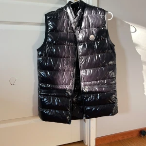 Svart dunväst från Moncler - Snygg svart dunväst från Moncler med glansig finish och quiltad design. Västen har ståkrage, två dragkedjefickor framtill och klassisk Moncler-logga på bröstet. Perfekt lager-på-lager-plagg för kyligare dagar. jag är en och 65 väger 45 och västen sitter lite Oversize på mig över midjan. se sista bilden hur den sitter.