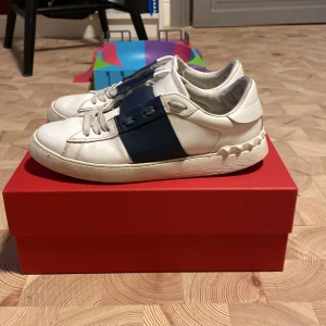 Valentino Garavani sneakers vit/blå - Snygga Valentino Garavani sneakers i vitt skinn med bred mörkblå rem över vristen och ikoniska nitar runt hälen. Klassisk rund tå, vita snören och platt sula. Perfekta för dig som vill ha en lyxig och stilren look med en cool twist. Priser kan diskuteras. Kan bytas mot marinblå maison margiela eller Louboutins inte svarta helst mörkblå med spika.