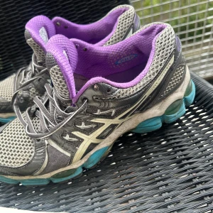 ASICS Gel Nimbus 14 sneakers grå/lila - ASICS Gel Nimbus 14 sneakers i grått med lila innerfoder och detaljer, samt turkosa inslag på sulan. Ovandel i mesh och syntet för bra andningsförmåga. Klassisk snörning och dämpad sula för extra komfort. Perfekta för dig som gillar sportig stil.