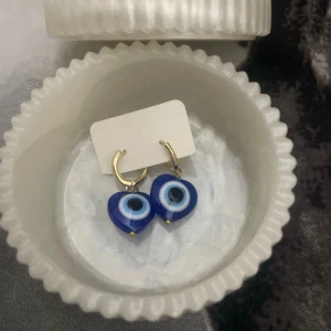 Blå evil eye örhängen med hjärtan/ 18 k guldpläterade hoops - Unika örhängen med blå evil eye-berlocker i hjärtform, hängande på små guldfärgade hoops. Perfekt statement-accessoar med coolt ögonmotiv och snygg kontrast mellan blått och guld. Köpta i Turkiet 