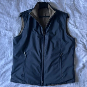 Blå och beige väst från Loro Piana - Snygg blå och beige vändbar väst från Loro Piana med Storm System-teknologi. Ena sidan är marinblå i polyester med dragkedja och fickor, andra sidan är beige i linne. Perfekt för lager-på-lager och stilren look. Hög krage och diskret broderad logga i nacken.