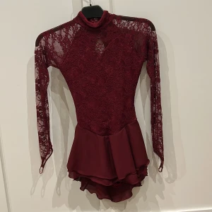 Vinröd konståkningsklännining med spetts och öppen rygg - Vinröd konståkningsklänningmed långa spetsärmar och glittrande detaljer. Klänningen har krage, omlottliknande öppen rygg med korsade band och volangig kjol i flera lager. Materialet är spets och polyester, vilket ger en lyxig och elegant känsla. Man får med ett matchande hår band.💕