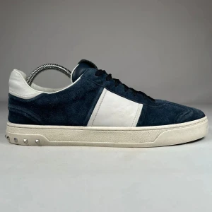 Valentino Flycrew - Snygga sneakers från Valentino Garavani i mörkblå mocka med vita detaljer. Skorna har en vit platt sula med alla nitar baktill och en klassisk rund tå. Perfekta för dig som vill ha en stilren men lyxig look. Dessa säljs inte längre i butik och inget og. Pris kan diskuteras