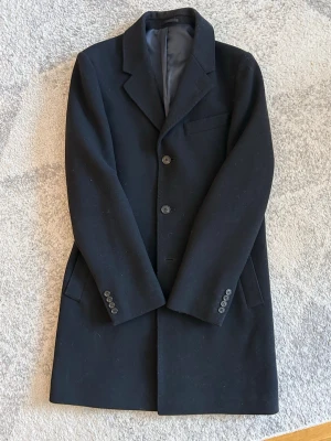 Svart rock från These Glory Days - Klassisk svart rock från These Glory Days i ullmix. Rak passform med tre knappar framtill, slät krage och fickor på sidorna. Fyra knappar vid ärmslutet ger en clean och stilren look. Inköpt på VOLT i Gbg. Nypris 2800kr. Hör av er vid intresse frågor!