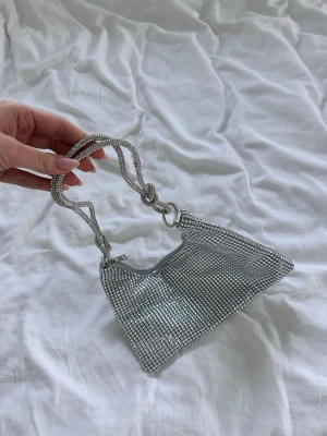 Glittrig silver axelväska - Säljer en glittrig axelväska i silver med ett snyggt rutmönster av små stenar. Perfekt för att lyfta din outfit med lite extra bling. I enkelt tunnt material från Amazon