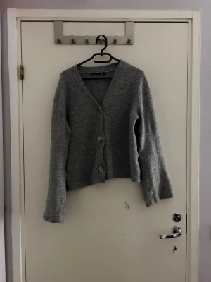 Grå stickad kofta med knappar - Mysig grå stickad kofta från lager 157 med v-ringning och tre stora knappar framtill. Koftan har lång ärm och en något loose passform, perfekt att slänga över en topp. Enkel och stilren design som funkar till det mesta.