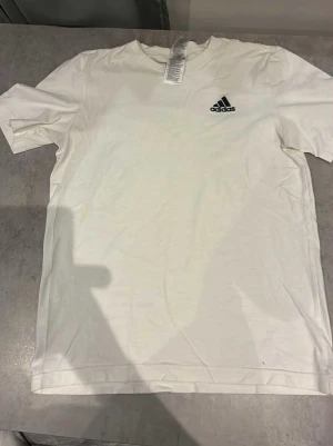 Vit Adidas t-shirt i bomull - Klassisk vit t-shirt från Adidas med svart broderad logga på bröstet. T-shirten har rund halsringning och korta ärmar. Tillverkad i mjuk bomull som känns skön mot huden. Perfekt till en avslappnad streetwear-look.