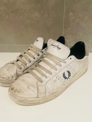 Fred perry skor - Klassiska vita sneakers från Fred Perry med svart logga på sidan och grå detaljer vid hälen. Skorna har snörning, rund tå och platt sula. Tillverkade i skinn och har en stilren, tidlös look som passar till det mesta.