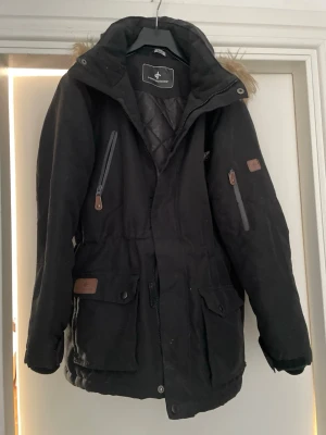 Svart parkas från Cross Sportswear - Säljer en svart parkas från Cross Sportswear med huva och fuskpälsdetalj. Jackan har flera fickor med både dragkedja och knappar, quiltat innerfoder och justerbar midja. Perfekt för kalla dagar och riktigt snygg till streetwear-stilen.