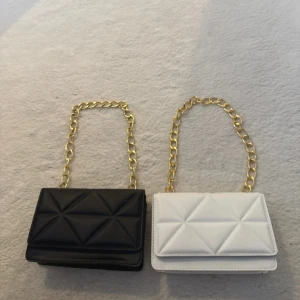 Svart och vit quiltad axelväska med kedja - Två stilrena axelväskor i svart och vitt med cool quiltad design och guldig kedja som axelrem. Väskorna har geometriskt mönster på framsidan och stängs med magnetlås. Perfekta för att lyfta din outfit med en trendig touch. Går att köpa bara en om man vill. En väska kostar 45 kr styck.