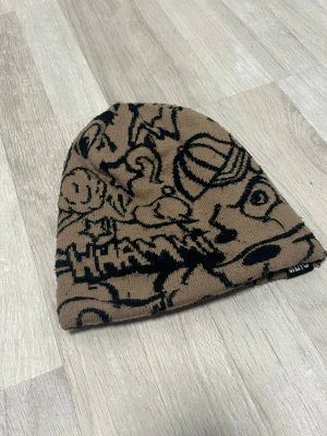 Brun mössa med svart grafiskt mönster - Cool brun mössa med svart grafiskt mönster och tecknade detaljer, inklusive texten 'OUCH' på ena sidan. Mössan är ribbstickad och har en avslappnad passform. Perfekt för dig som vill sticka ut med en unik accessoar.  brand ALAB