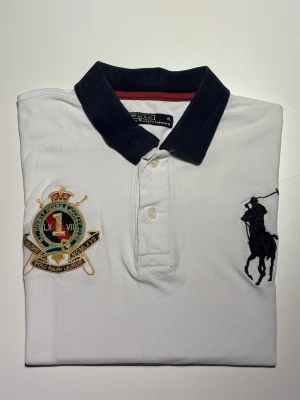 Ralph Lauren piké - Crest Logo - Klassisk Polo Ralph Lauren piké polo i vit färg med exklusivt broderat crest-emblem och big pony. Tidlös design med högkvalitativ pikéväv och ikonisk passform. I mycket fint skick, sparsamt använd. Ett stilrent och eftertraktat plagg som passar både vardag och mer uppklädda tillfällen  Storleken är skriven som XL men passar mer som Medium.