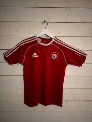 Röd Bayern München Adidas tröja - Säljer en röd Bayern München tröja från Adidas med vita detaljer och logga på både fram- och baksidan. Tröjan har rund hals och korta ärmar, samt tre vita ränder längs axlarna. Perfekt för dig som älskar fotboll och vill representera ditt favoritlag.