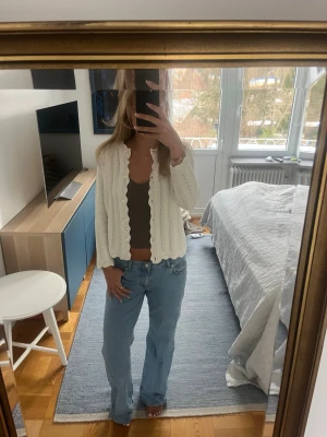 Vit stickad kofta med vågig kant - Säljer en söt vit stickad kofta med vågig kant och dekorativa knappar framtill. Koftan har lång ärm och ett luftigt, hålmönstrat stickat material som ger en fräsch och trendig look. Perfekt att slänga över ett linne eller t-shirt!