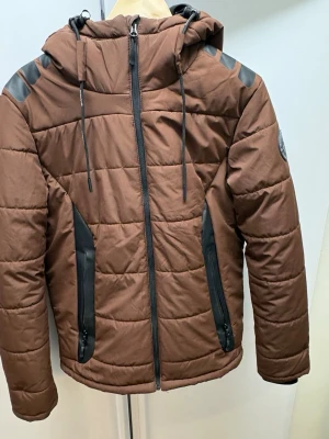 Brun pufferjacka från Bluewear - Säljer en brun pufferjacka från Bluewear med svarta detaljer och huva. Jackan har dragkedja framtill, två stora fickor med dragkedja och coola svarta markeringar på axlar och huva. Perfekt för kalla dagar och riktigt skön passform. Passar som M 