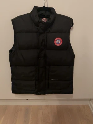 Svart dunväst från Canada Goose - Svart dunväst från Canada Goose med klassisk logga på bröstet. Västen har två stora fickor framtill, hög krage och dragkedja. Perfekt för kalla dagar och har en stilren, quiltad design. Snygg och funktionell för dig som gillar streetwear och outdoor. Storlek L, passar även M.
