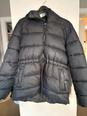 Svart pufferjacka från Everest dam 36  - Säljer en svart pufferjacka från Everest i storlek 36 (s/m).  Ävänds en säsong, jättebra skick. Jackan har huva med dragsko och två stora fickor med dragkedja. Midjan kan justeras med snörning för bättre passform. Jackan är mjuk och riktigt skön att bära. Säljer pga vinter garderobsrensning. 