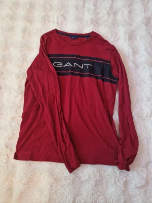 Röd tunn långärmad tröja från GANT - Säljer en röd långärmad tröja från GANT med svart och vit logga över bröstet. Toppen har en klassisk rund halsringning och är gjord i mjuk bomull. Perfekt för en avslappnad och stilren look.
