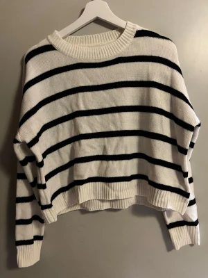 Randig stickad tröja i vitt och svart - Säljer en snygg stickad tröja med svartvita ränder. Tröjan har en relaxed fit med ribbade muddar vid hals, ärmslut och nederkant. Perfekt för dig som gillar en enkel men trendig look. Passar till både jeans och kjol.