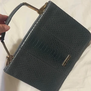 Grå handväska i ormmönstrat skinn - Säljer en grå handväska med ormmönstrad struktur och guldfärgade metalldetaljer. Väskan har ett stilrent handtag och rektangulär form, perfekt för dig som gillar en edgy men klassisk look. Märkesplakett framtill för extra lyxig känsla.