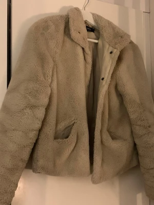 Beige teddyjacka från ONLY - Supermjuk och fluffig teddyjacka i beige från ONLY. Jackan har klassisk krage, två fickor framtill och stängs med tryckknappar. 