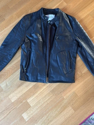 Svart skinnjacka från Glory Days - Säljer en svart skinnjacka från Glory Days med stilren design. Jackan har ståkrage med tryckknapp, tre dragkedjeförsedda fickor framtill och detaljerade sömmar på axlarna. Passformen är normal och materialet är mjukt skinn. Är 184 cm väger 83 kg, satt lite tajt för mig och var lite kort men perfekt för dig i storlek s/m och lite mindre byggd. 