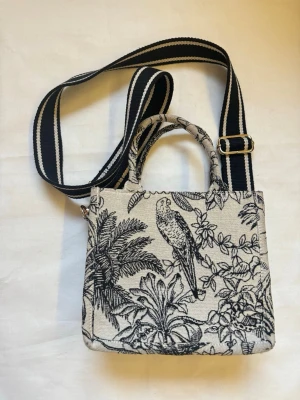 Beige och svart axelremväska med fågelmönster från H&M - Produktbeskrivning •Snygg svart och vit axelremväska. •Köpt från H&M. •Med broderat mönster av djur och växter. •Väskan har både handtag och en bred, justerbar axelrem i svart och vitt. •Perfekt både till vardag och till jobb. •Trendig “clean girl / classy” stil •Har fläckar från blyertspenna på insidan - går nog bort med tvätt. •Väskan har inte en dragkedja utan är öppen. •Väskan har en innerficka med dragkedja.   Mer information  •Skickas inom 24h! •Vid fler frågor eller om du vill ha fler bil